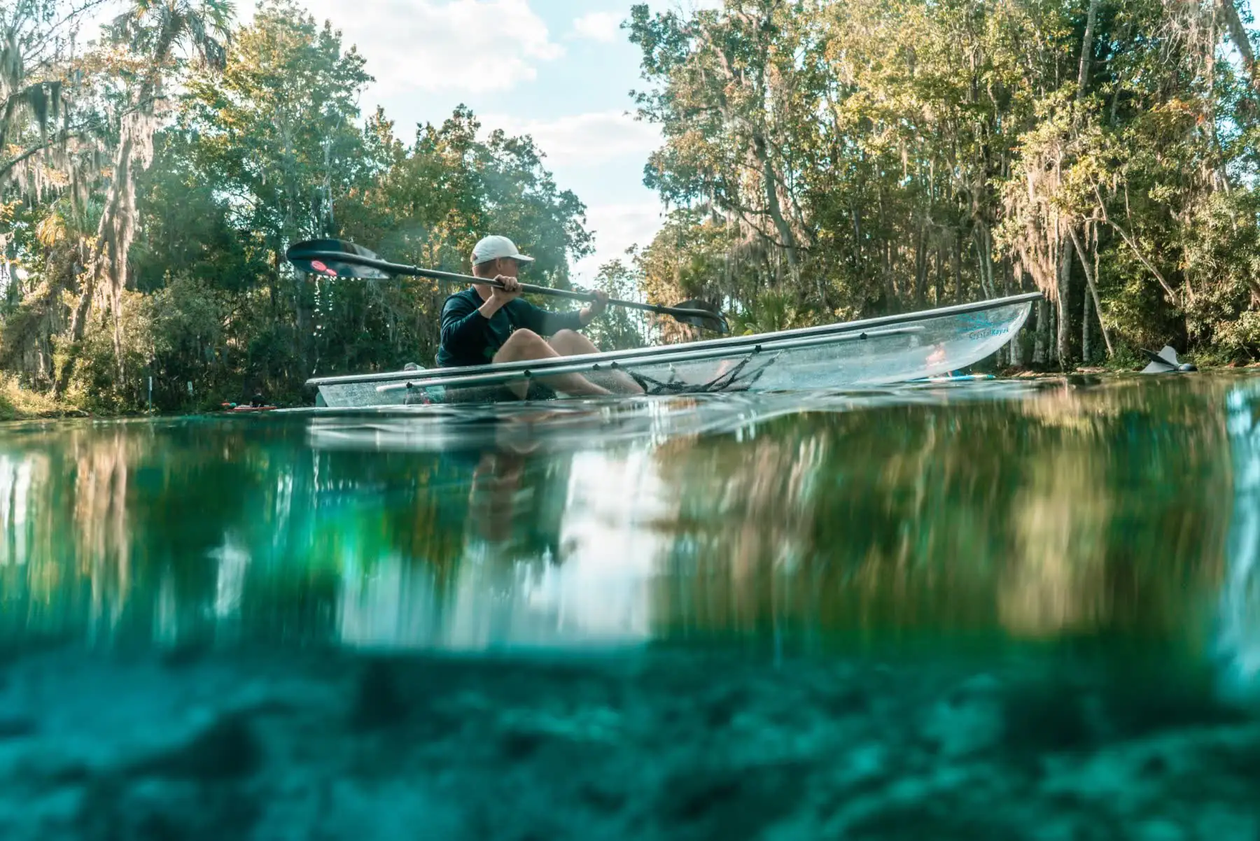 Half-underwater view of a clear kayak, trees above and spring floor below