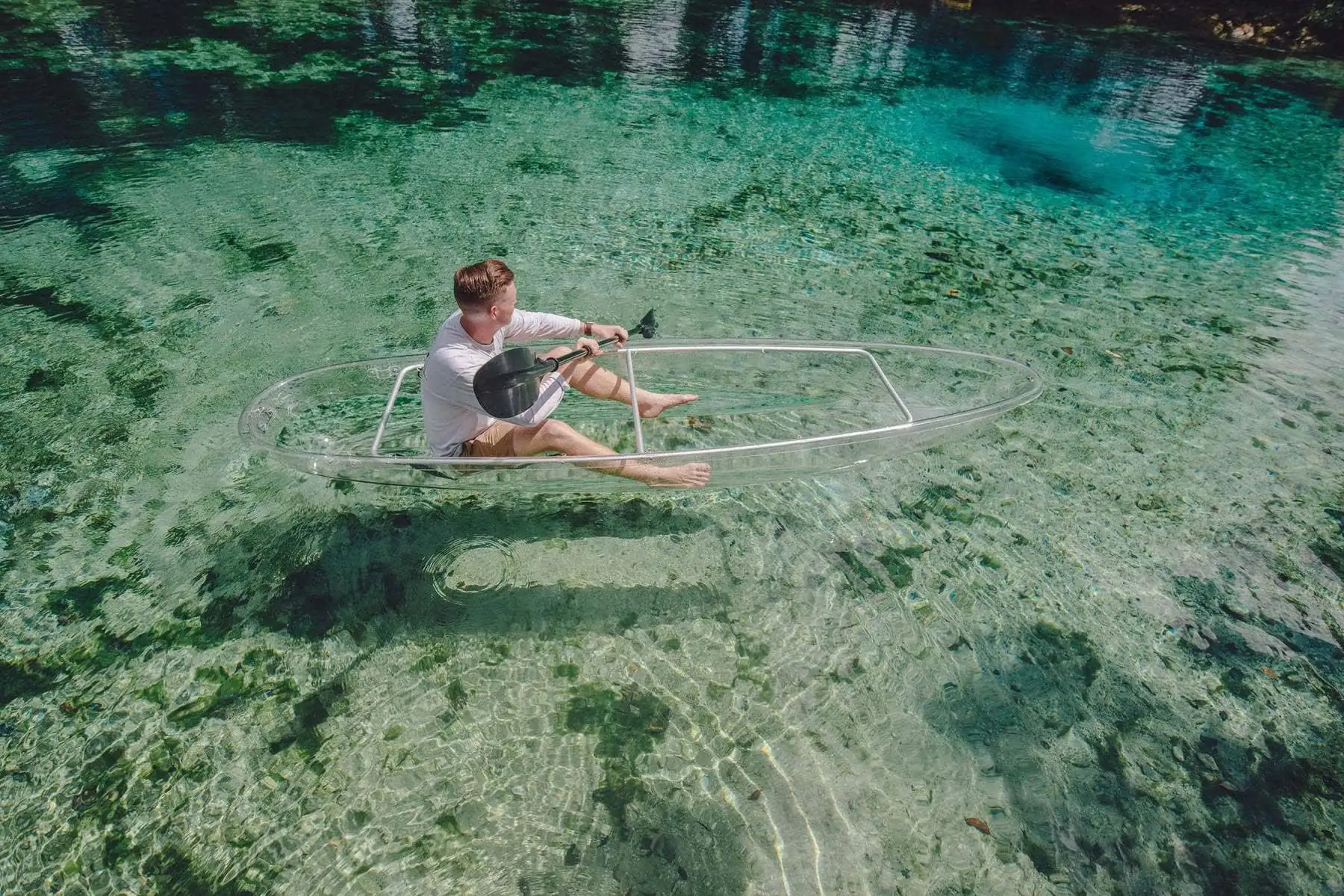 A paddler sits in a fully transparent kayak floating above green seagrass in clear spring water